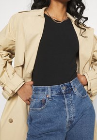 Abrigo trench beige con cinturón, blusa negra ajustada y jeans de tiro alto en azul con costuras visibles y remaches de cobre.