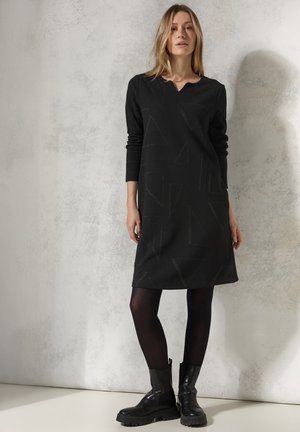 Robe noire à manches longues avec des motifs géométriques, décolleté en V et en tissu doux ; associée à des bottes noires à talons épais et des collants opaques.
