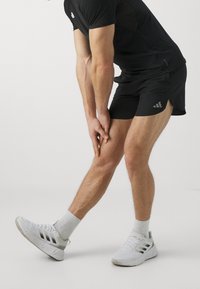 Svart sporttröja och shorts, tillsammans med vita sneakers. Shorts har sidofickor; tröjan har en figurnära design. Slät textur.