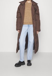 Cappotto puffo marrone con colletto grande, maglione in maglione beige, jeans azzurri chiari e scarpe neri con suola spessa, posizionato su uno sfondo neutro.