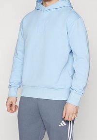 Uomo che indossa un hoodie azzurro chiaro e pantaloni da jogging grigi Adidas con strisce bianche, in piedi contro uno sfondo neutro e semplice.