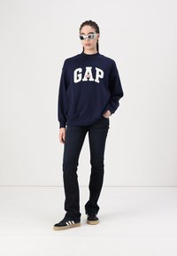 Navy blue sweatshirt with "GAP" logo, paired with dark jeans and black sneakers featuring white stripes and gum sole, accessorized with sunglasses.