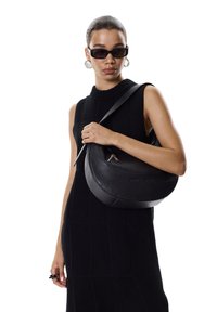 Sac à main en cuir noir avec une forme incurvée et une bandoulière réglable ; dispose d'une fermeture éclair et d'un détail de logo sur le devant.