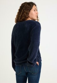 Laivansinisestä fleece-neuleesta, jossa on pehmeä pinta, laskettu olkalinja ja pitkät hihat; yhdistetty tiukkoihin sinisiin farkkuihin. Takaview esitetty.