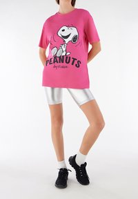 Maglietta di cotone rosa con grafica nera di Snoopy e il testo "PEANUTS by Schulz"; abbinata a pantaloni ciclistici argentati lucidi e sneakers nere.