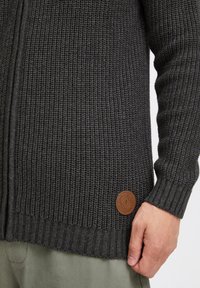Strickcardigan in Dunkelgrau mit gerippten Bündchen und Saum. Hat ein rundes Lederlabel mit eingeprägtem Logo auf der unteren linken Seite.
