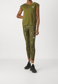 Olivgrön t-shirt med korta ärmar och en logotyp, kombinerad med mönstrade olivgröna leggings. Modellen bär svarta träningsskor med vita strumpor.