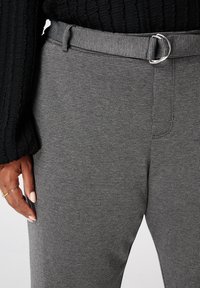 Grijs gemusterde broeken van een gestructureerde stof met een riemlus en een zilveren gesp. Close-up die een deel van een hand nabij de tailleband toont.