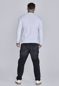 Fehér hosszú ujjú ing, sötét farmernadrággal párosítva, laza szabású, két hátsó zsebbel. Fehér tornacipővel egészül ki a megjelenés.