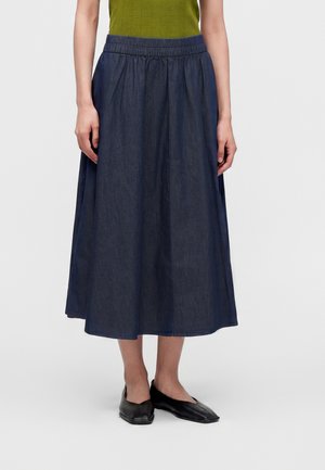 Femme portant une jupe midi bleu foncé avec une taille élastique, un haut vert et des chaussures plates noires, debout devant un fond clair uni.
