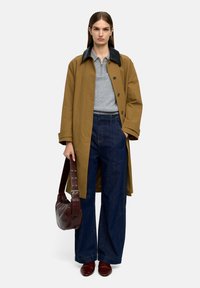 Brązowy płaszcz z czarnym kołnierzem, szary polo, szerokie ciemnoniebieskie dżinsy, burgundowa torebka i pasujące buty; gładkie faktury, detale z guzikami.