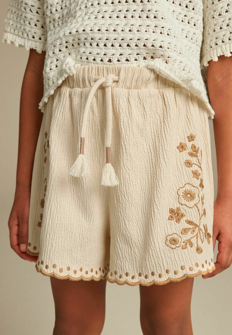 Shorts beige texturés avec taille élastique, cordons avec pompons à l'avant, ourlet festonné et broderie florale marron, portés avec un haut en crochet crème.