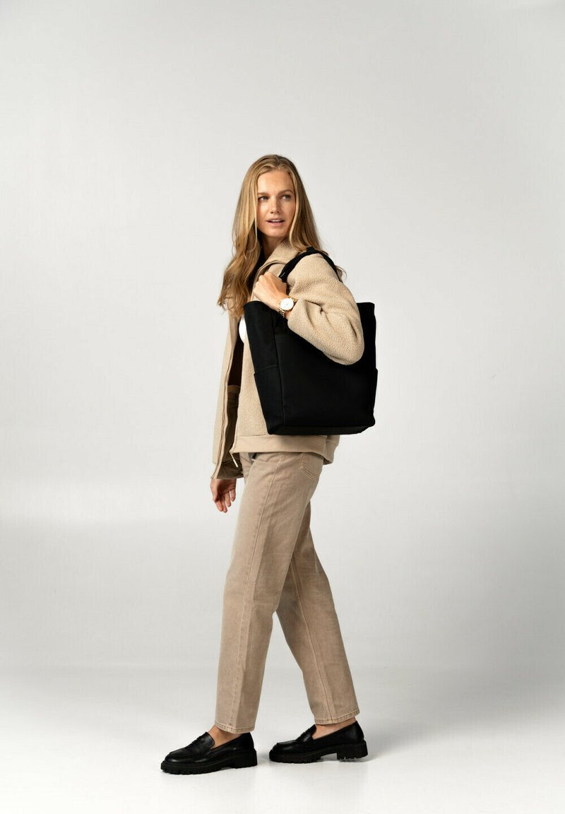 Borsa tote nera con manici robusti, forma rettangolare e superficie liscia. Indossata da una persona in un outfit beige su uno sfondo neutro.