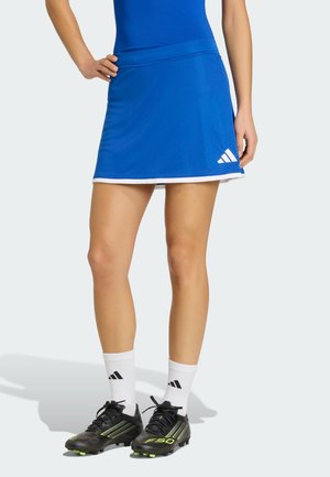 Mujer con falda deportiva azul con ribete blanco, calcetines blancos y tacos negros, de pie con una mano en la cadera.