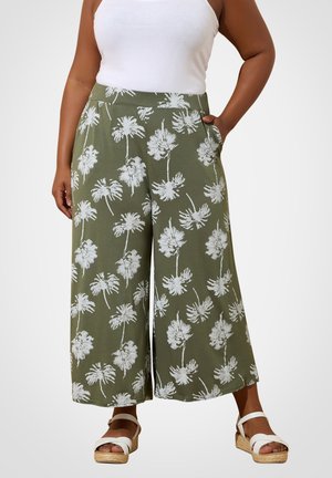 Vrouw draagt olijfgroene broek met wijde pijpen en witte palmboomprints, gecombineerd met een wit mouwloos topje en witte sleehakken.
