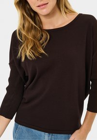 Femme aux longs cheveux blonds portant un haut en maille côtelée marron foncé à manches mi-coude et un jean bleu sur fond blanc.