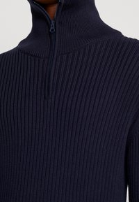 Marinblå ribbad polotröja med halv dragkedja. Har en texturerad stickad design och en figurnära silhuett. Materialet verkar mjukt och tjockt.