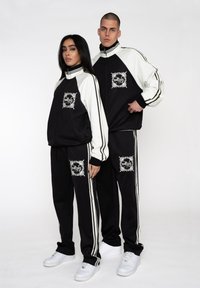 Sort-hvide tracksuits med strikstruktur, jakker med lynlås og stribede detaljer. Omfatter grafiske udsmykninger på brystet og låret.