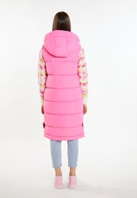Langer pinker Steppwesten mit Kapuze, mit horizontalen Nähten und seitlichen Schlitzen. Trägt sie über einem farbenfrohen gemusterten Pullover und hellen Jeans.