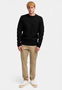 Schwarzer Strickpullover mit geometrischen Mustern, Rundhalsausschnitt und enger Passform, kombiniert mit beigen Hosen und weißen Turnschuhen.