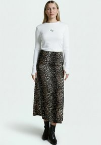 Weißes Langarmoberteil mit kleinem schwarzem Logo, kombiniert mit einem Maxirock in schwarz-beigem Leopardenmuster. Es werden schwarze Stiefeletten getragen.