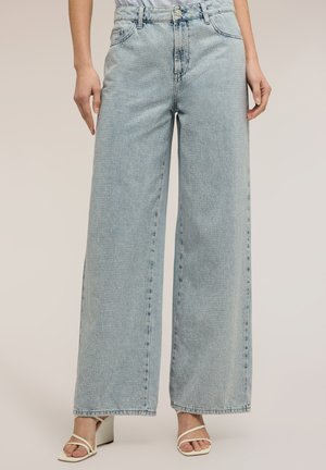 Jeans a vita alta color azzurro chiaro con gamba larga e sottili puntini testurizzati, abbinati a sandali bianchi con tacco e cinturini su uno sfondo neutro.