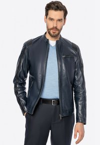 Giubbotto di pelle blu navy con colletto rialzato, chiusura con zip e spalle a costine. Texture liscia e design aderente; indossato sopra una camicia azzurro chiaro.