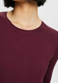 Viininpunainen ribattu neule, jossa on pyöreä pääntie. Kangas esittää hienoja pystysuoria viivoja, jotka luovat teksturoidun ulkonäön.