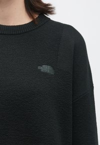 Κοντινή λήψη ενός ατόμου που φοράει ένα μαύρο υφασμάτινο πουλόβερ με ριγωτό γιακά και ένα μικρό κεντημένο λογότυπο The North Face στο στήθος.