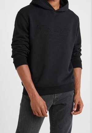 Zwarte hoodie van zacht materiaal, met een gestructureerd embleem op de voorkant, geribbelde manchetten en een voorzak in kangoeroevorm.