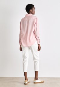 Camicia di cotone rosa chiaro con cucitura posteriore, vestibilità comoda e orlo arrotondato, abbinata a jeans bianchi a vita alta e scarpe con dettagli argentati.