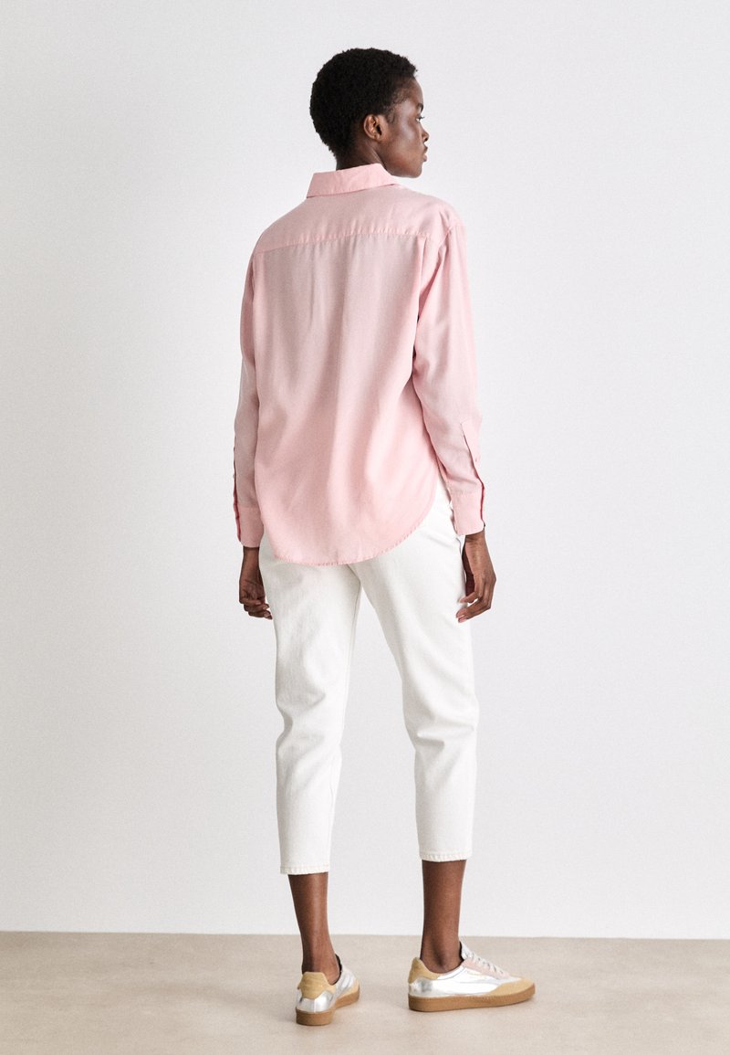 Chemise en coton rose clair avec couture au dos, coupe décontractée et ourlet arrondi, associée à un jean blanc coupe cropped et des chaussures aux accents argentés.
