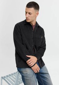 Camisa de pana negra con botones en la parte delantera, cuello y un solo bolsillo en el pecho; llevada sobre una camisa de color burdeos y combinada con unos vaqueros azules desgastados.
