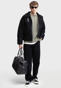 Black shearling jacket with leather accents, green cable-knit sweater, black pants, and black sneakers; holding a leather duffel bag.