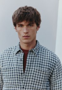 Koszula w kratę, button-up, w niebiesko-białym kolorze, o miękkiej teksturze i jednej kieszeni na piersi, noszona na burgundowym t-shircie z okrągłym dekoltem.