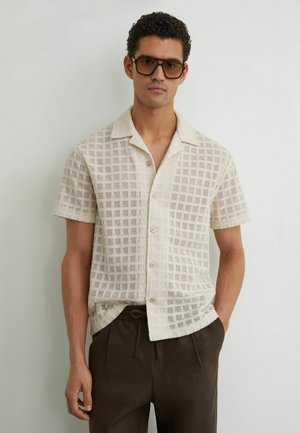 Mees, kes kannab üle suurusega pruune päikeseprille, beežikat lühikese varrukaga ruudulise mustriga nööbitavat särki ja tumepruune nöörikutsega pükse.