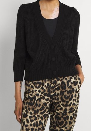 Femme portant un cardigan noir en maille boutonné sur un haut noir, associé à un pantalon taille haute imprimé léopard, debout les mains dans les poches.