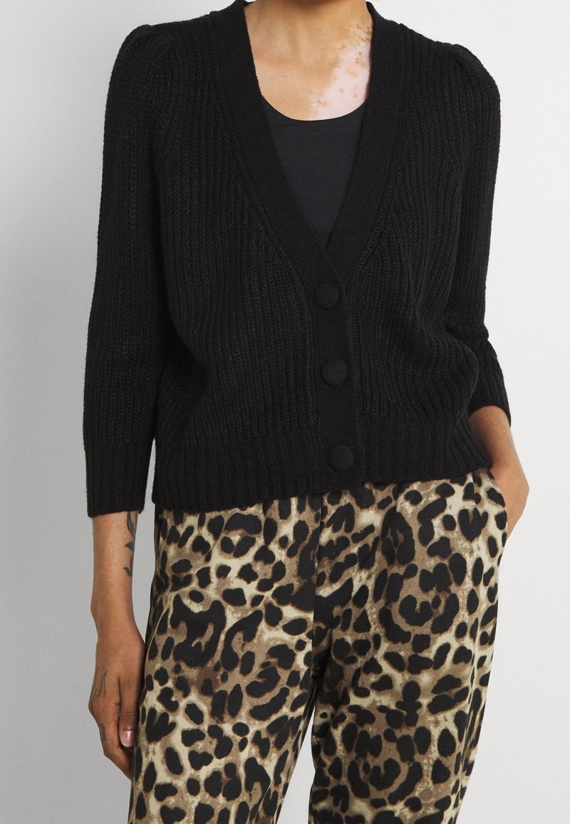 Femme portant un cardigan noir en maille boutonné sur un haut noir, associé à un pantalon taille haute imprimé léopard, debout les mains dans les poches.