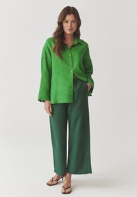Groene linnen overhemd met een knoopsluiting aan de voorkant, gecombineerd met losse groene broeken. Zachte textuur en relaxte pasvorm, aangevuld met platte sandalen.