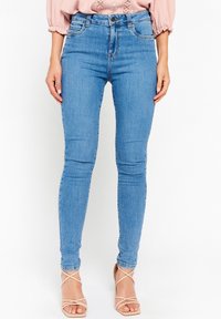 Kvinna som bär ljusblå slim fit-jeans och nakna sandals med hög klack, stående mot en vit bakgrund.
