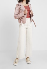 Person, der eine blassrosa Bikerjacke, ein rostfarbenes, knöpfbares Top, hochtaillierte, weiße, weite Hosen und beigefarbene Sneakers vor einem weißen Hintergrund trägt.