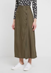 Olivgrüner Midirock mit einem Frontschlitz und fünf Knöpfen an der Seite. Hergestellt aus glattem Stoff, kombiniert mit weißen Sneakern.