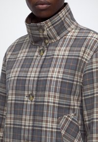 Gros plan d'une personne portant une veste à carreaux gris, marron et blanc avec boutons, un col montant et une poche sur la poitrine.
