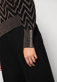 Svart och metallisk stickad tröja med zigzag-mönster och ribbade ärmslut som har dekorativa knappar. Slät textur med en figurnära silhuett.