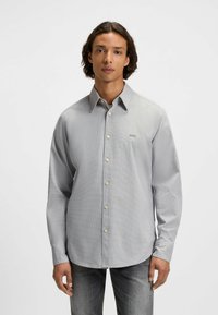 Helle graue Langarmknopfhemd mit strukturiertem Muster und Logo auf der linken Brust, kombiniert mit dunklen Jeans. Glattes Baumwollmaterial.