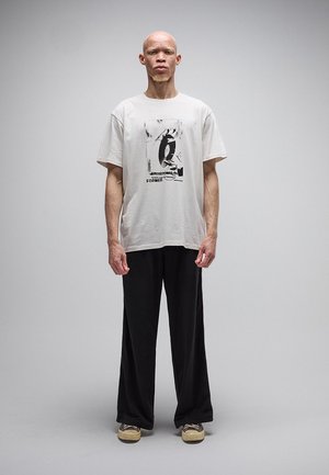 Mand med albinisme stående, iført en hvid grafisk t-shirt, sorte vidde ben bukser og beige sneakers mod en ensfarvet grå baggrund.