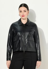 Musta nahkatakki, jossa on kaulus, etuvetoketju, kaksi paikkataskua ja sileä pinta. Suunniteltu istuvaan siluettiin.