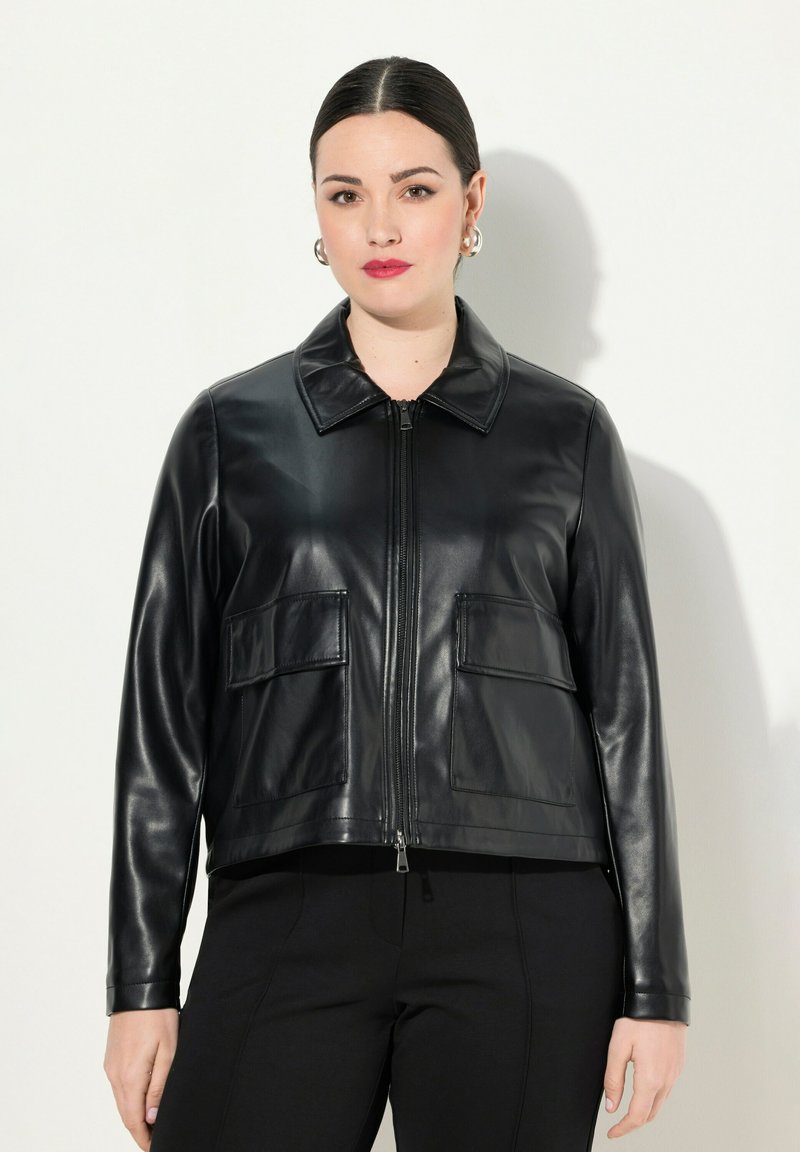 Musta nahkatakki, jossa on kaulus, etuvetoketju, kaksi paikkataskua ja sileä pinta. Suunniteltu istuvaan siluettiin.