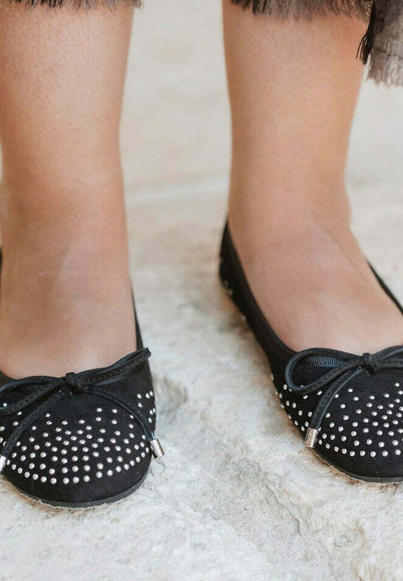 Pies que llevan bailarinas negras adornadas con pequeños remaches plateados y atadas con delgadas cintas negras, de pie sobre una superficie texturizada clara.