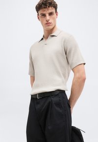 Νέος άνδρας που φοράει μπεζ πλεκτό πόλο με κοντά μανίκια και φαρδιά μαύρα παντελόνια με μαύρη ζώνη, στέκεται μπροστά σε απλό ανοιχτόφωτο φόντο.